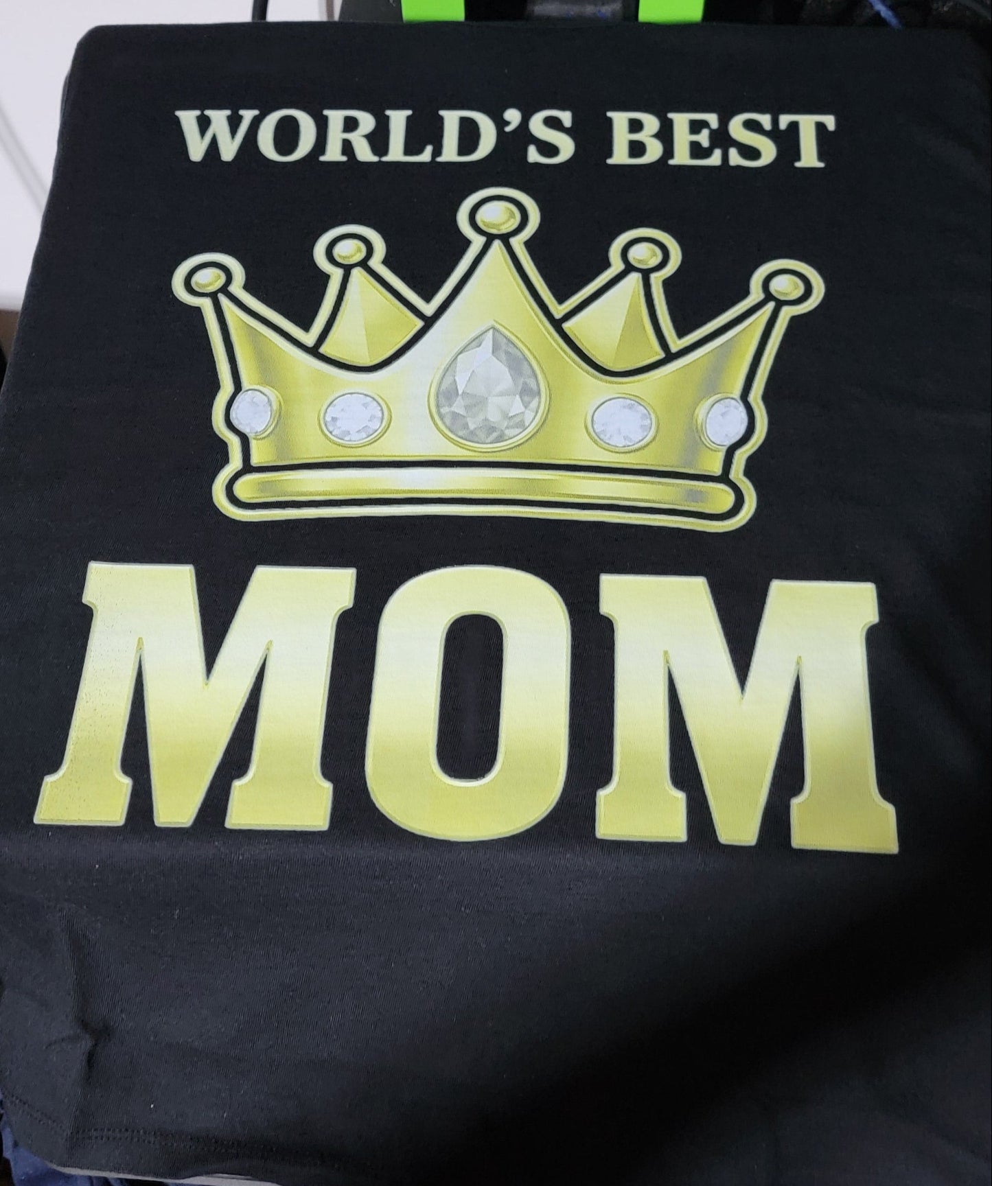 Black shirt with gold crown and 'World's Best Mom' text worn by a person, with shoes and a bag in the background.